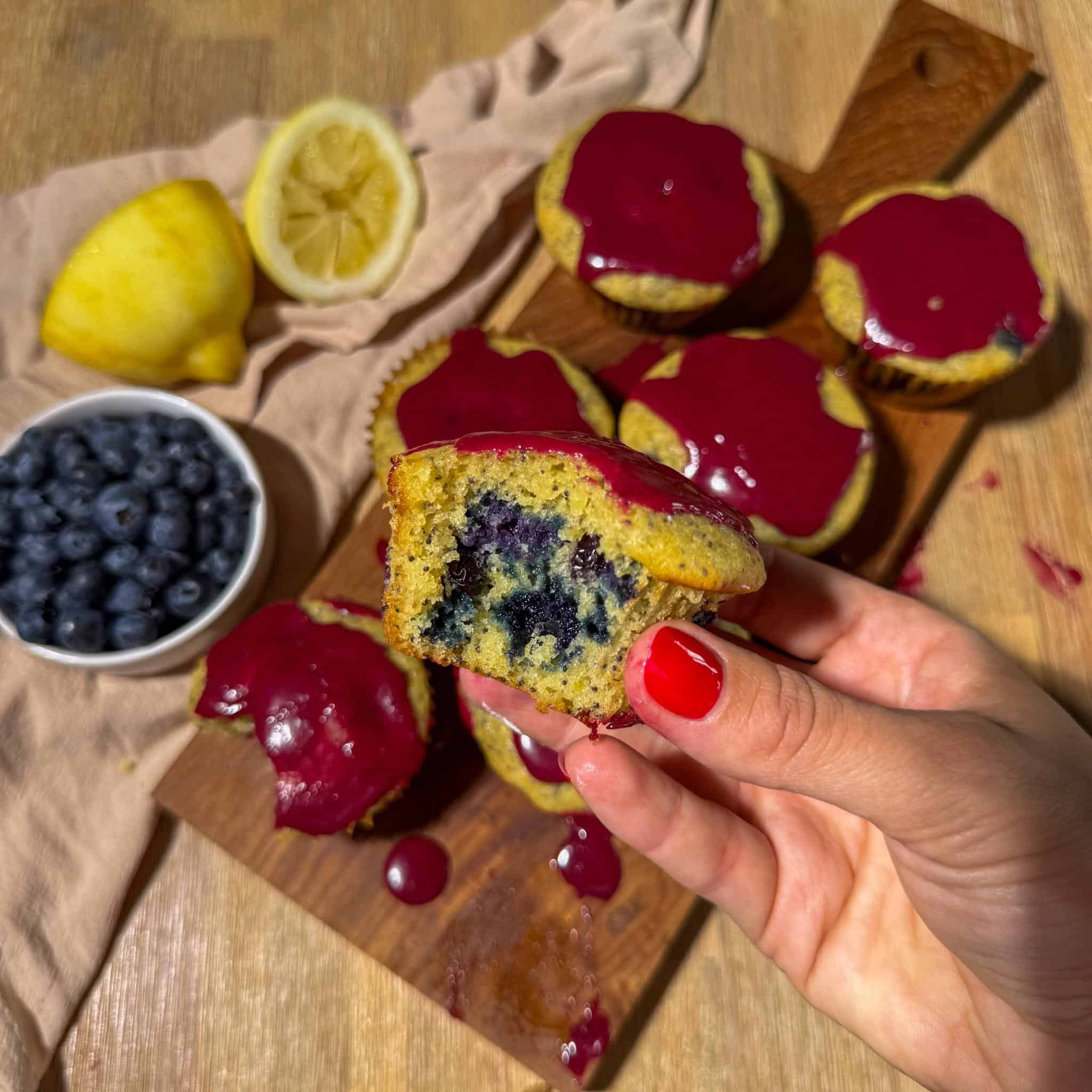 Blueberry Lemon Poppyseed Muffins 8 hand holding one blueberry lemon poppyseed muffin with a bite taken out exposing the blueberry-filled center, above a board of glazed muffins