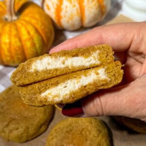 Pumpkin Chai Cheesecake-Stuffed Cookies 7 Gooey pumpkin chai cookies with a cheesecake filling cut in the center and held n a hand, fall themed snacks with pumpkins in the background, perfect for autumn treats