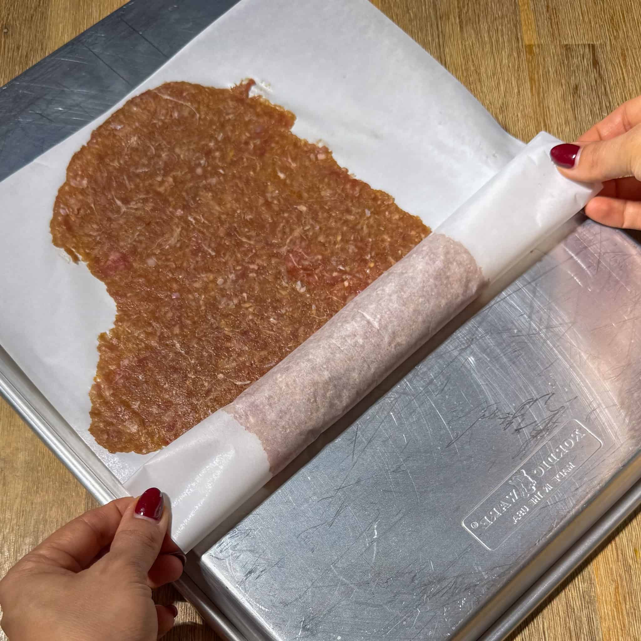 Two hands rolling up the parchment paper that has the meat mixture laid flat.