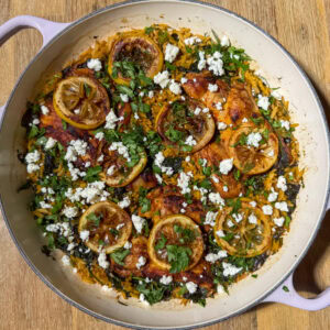 Round pan full of baked lemon chicken thighs and orzo topped with feta and herbs and caramelized lemons