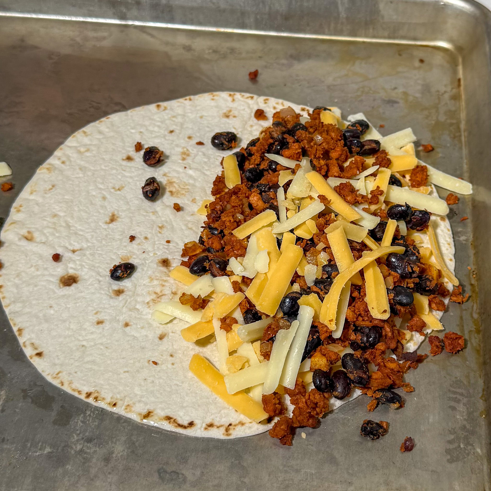 Uncooked tortilla on a sheet tray, with a layer of cheese then the black bean soyrizo mixture, then more cheese, before baked.