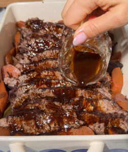 Red wine braised brisket in a serving platter surrounded by vegetables with a hand pouring sauce over the meat