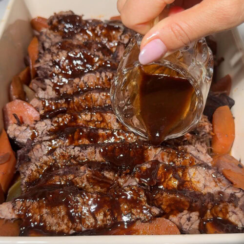 Red Wine Braised Brisket 11 Red wine braised brisket in a serving platter surrounded by vegetables with a hand pouring sauce over the meat